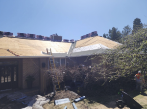 A single-story brick house undergoing a roof replacement, with exposed plywood decking, stacks of new shingles along the ridge, a yellow ladder, and a worker in an orange safety vest, set against a clear blue sky.