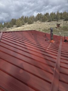 A stainless steel chimney pipe with an orange flashing boot being installed on a red corrugated metal roof, with tools scattered nearby and a rural landscape of trees and dry grassland in the background
