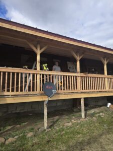 A rotated photo showing a wooden deck or porch construction project with fresh lumber framing, a DISH satellite dish mounted on a pole, power tools including a Ryobi nailer and DeWalt battery, a bag of GAF TigerPaw roofing material, and a person in a gray shirt and cap working on the structure
