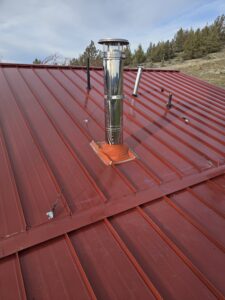 A stainless steel chimney pipe with a guy-wire bracing system installed through an orange flashing boot on a red corrugated metal roof, with trees, mountains, and blue sky in the background.