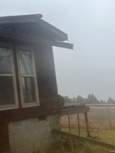 Corner of a dark wood-sided house with white-trimmed windows and a concrete block foundation, showing storm-damaged roofing hanging off, set against a foggy field and tree line under an overcast sky