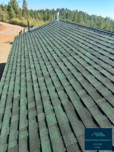 Newly installed dark green architectural asphalt shingle roof photographed along the hip ridge, with a metal vent pipe and stove pipe near the peak, pine forest and blue sky in the background, and a Wright's Roofing & Construction inc logo watermark in the lower right corner.