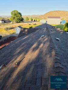 Aerial view along the ridge of a freshly installed brown architectural asphalt shingle roof, showing ridge cap shingles, metal box vents and pipe boots, with a rural ranch landscape, cattle pasture, barns and hills in the background; Wright's Roofing & Construction Inc watermark in the corner.
