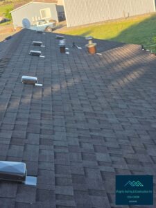 Aerial view of a freshly installed dark gray asphalt shingle roof featuring multiple black box vents, pipe flashings, and a metal chimney cap, with a metal barn, farm truck, and green fields in the background. Wright's Roofing & Construction Inc logo overlay in the lower right.