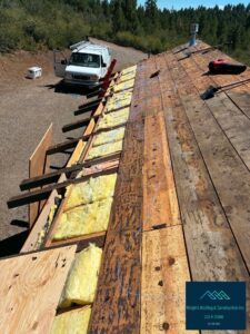 Aerial view of a roof replacement in progress, showing stripped decking boards, exposed rafter tails with yellow fiberglass insulation, new plywood sheathing, a red ladder, and a white work van parked on gravel below with forested hills in the background.