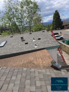 Aerial view of a residential roof mid-replacement, showing gray asphalt shingles with vents, pipe boots and a skylight alongside a stripped lower section, with an orange ladder on the deck and mountain scenery in the background. Watermarked Wright's Roofing & Construction inc, CCB #241098.