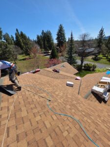 A roofing worker in a safety harness kneels on a large tan asphalt shingle roof, surrounded by air hose, rope, nail gun and open shingle boxes, with pine trees and a sunny blue sky in the background.