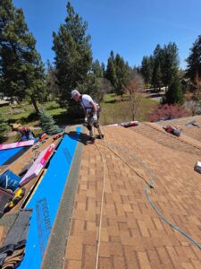A roofing worker in a safety harness and cap stands on a shingled ridge, using a nail gun while blue synthetic underlayment, tools, and shingle bundles sit on the exposed deck beside him under a clear blue sky.