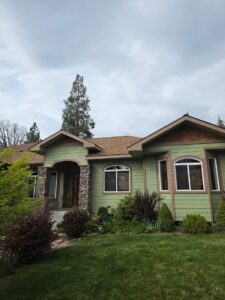 A single-story sage green craftsman-style home with brown trim, stone-veneer porch columns, arched entry, and shingled gables, framed by manicured lawn and shrubs under an overcast sky.